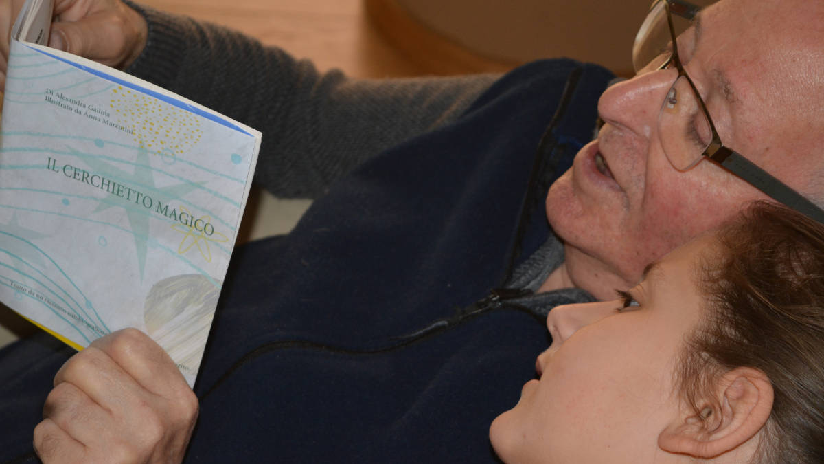 Una bambina seduta assieme al nonno legge il racconto illustrato sulla sordità Il Cerchietto Magico